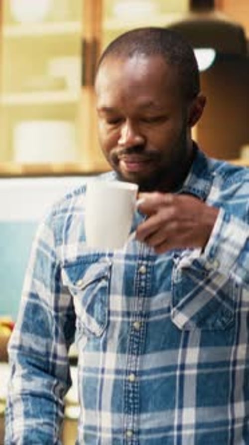 Vertikales Videoporträt eines fröhlichen jungen Mannes, der eine Tasse frischen Kaffee an der Theke hält