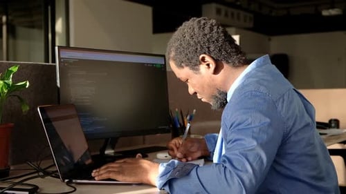 Homem negro escrevendo linhas de código em um PC de mesa com dois monitores e um laptop ao lado em um escritório elegante
