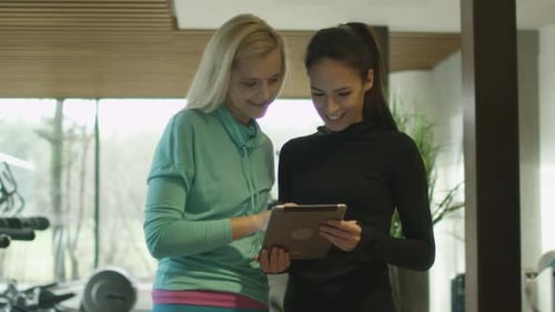 Two beautiful athletic women are using a tablet next to treadmills in a sport gym.