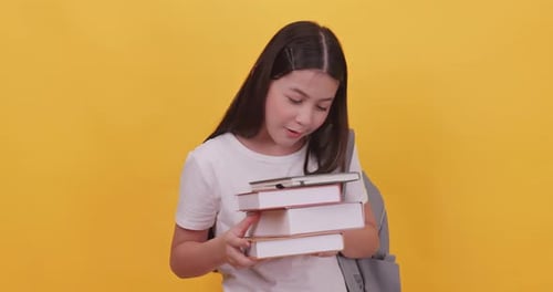 Cute school girl is carrying a school bag and holding a textbook. Isolated on yellow background