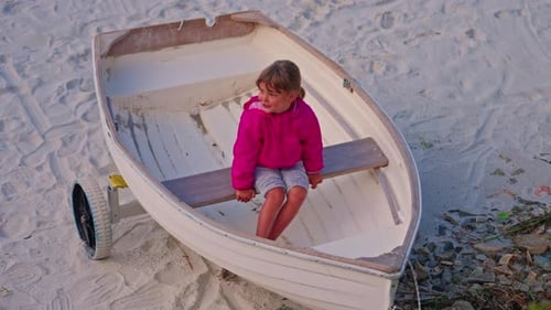 Kleines schönes Mädchen in rosa Jacke sitzt im Fischerboot am Sandstrand