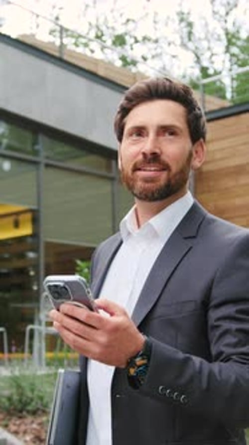 Confident Businessman in a Formal Suit Holding a Laptop and Using Smartphone for Online Messaging or