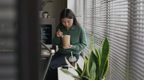 Woman Eats Noodles at Desk by Window