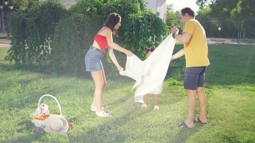 The Family Spreads a White Blanket on the Green Grass Setting Up Their Picnic Spot Under the Sunny