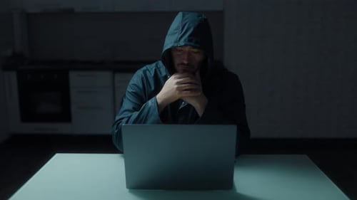 Man in hood types on laptop in dark room