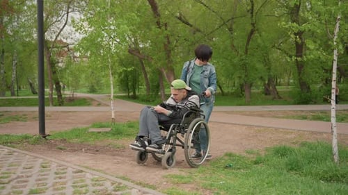 Person Using Wheelchair Being Assisted Along Park Trail With Nature Man Seated In Wheelchair Is