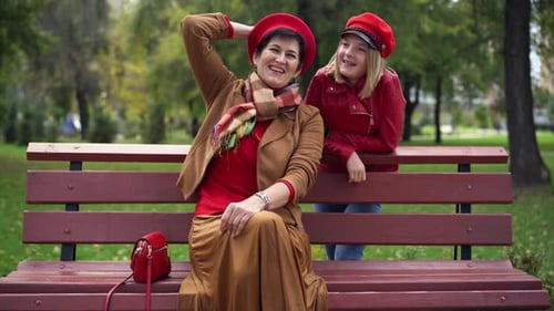 Joyful Senior Woman Sitting on Park Alley Talking with Teen Girl Leaning on Bench Smiling Positive