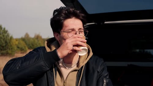 Close-up shot of a guy sitting in the trunk of an SUV in the steppe and drinking tea from thermos