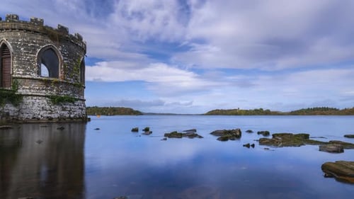 Timelapse of a medieval tower on lake with shoreline rocks in the foreground and medieval castle in