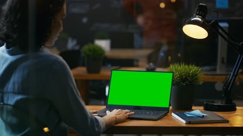 Female Office Worker at Her Desk Works on a Laptop with Mock-up Green Screen. Over the Shoulder Foo