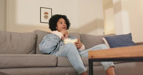 Mid adult African American man reacting to suspense show by scooping popcorn and laughing on couch