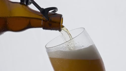 Brown beer bottle fills glass goblet with cold lager stream in super slow motion
