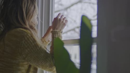 Woman Standing By Window Inside Home Looking Out
