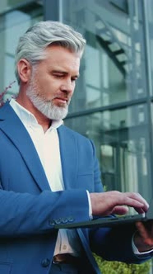 Businessman in White Shirt Sitting on Bench Working on Laptop Outdoors Near Modern Glass Building