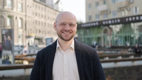 A Man in a Blue Suit and Glasses Looks Into the Camera and Smiles on the Street in the City