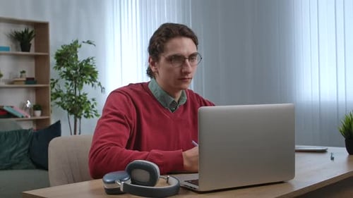 Young Adult Man Spending Time at Home Man Student Sits at Desk Works on Laptop Talking Online By