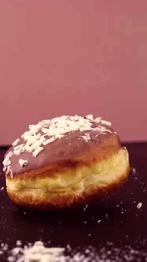 A Berliner donut with a glossy glaze and white shavings is dropped in slow motion. The fluffy dough