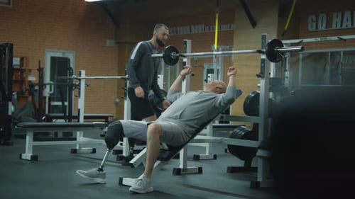 Athlete with Prosthetic Leg Trains with Barbell in Gym with Personal Trainer