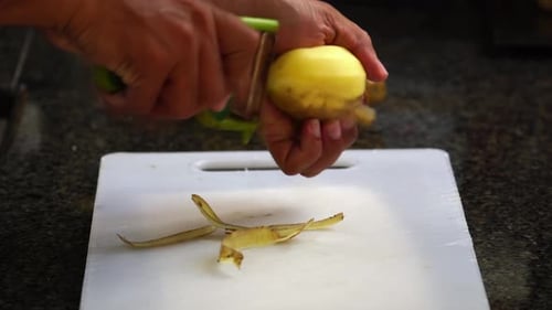 Middle aged brown skin man peeling potatoes with peeler on a white chopping board at home