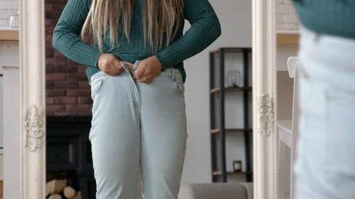 Woman struggles to button light jeans in mirror