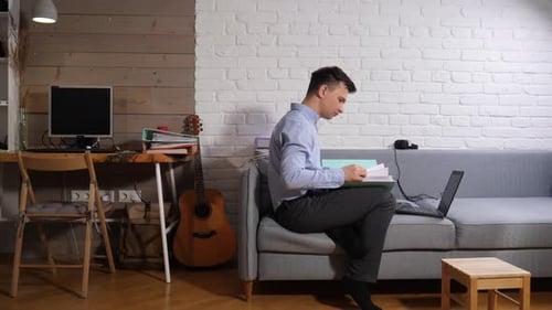 Junger Mann auf einem Sofa in einer modernen Wohnung, der remote von seinem Heimbüro aus arbeitet und sich auf den Laptop konzentriert
