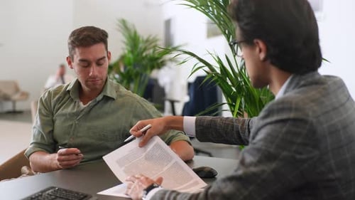 Portrait of Cheerful Young Man Client Reading and Signing Contract with Professional Male Realtor
