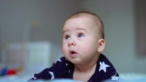 Cute baby lies on bed on his stomach. Beautiful infant child looks up with curiosity. Close up.
