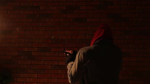Criminal man in hood with gun near brick building outdoors, back view