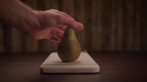 A Hand is Gracefully Holding a Ripe Pear Positioned Above a Cutting Board Placed on a Table