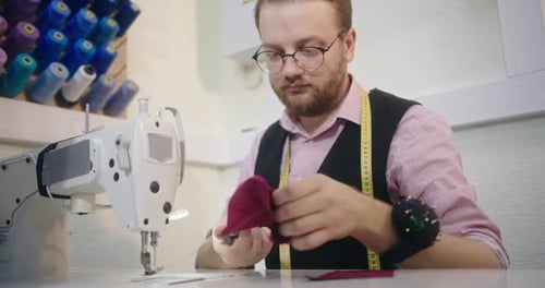 A Man with Glasses a Designer Who Sews Clothes Sitting at a Table in His Workshop Sews on a Sewing