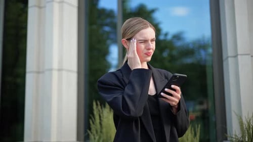 Woman Checks Phone, Rubs Temple in City Environment