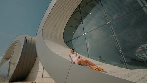 Stylish Model In White Swimsuit Lies On Background Of Modern Oval Building
