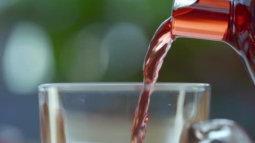 Close up pouring red herbal tea out of transparent tea pot into glass cup, green nature background.