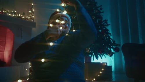 Happy Young Man Plays With Christmas Lights At Night Near The Tree In The Room