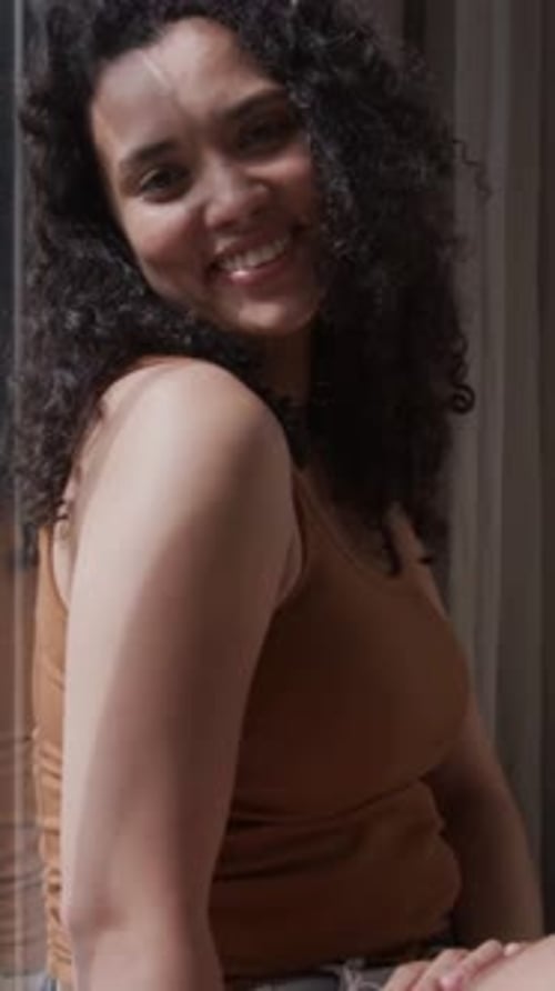 Vidéo verticale : Une femme souriante détourne le regard en baissant le regard par une fenêtre drapée alors que la lumière du soleil traverse le visage