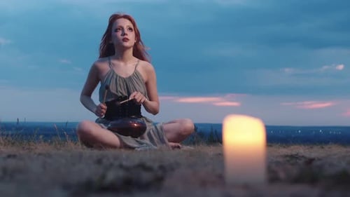A Redhaired Girl is Playing a Glucophone Outdoors Sitting on the Grass in the Candlelight