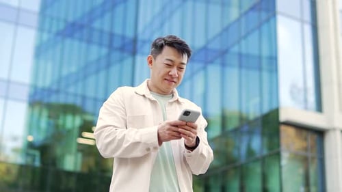 Happy asian man uses a smartphone while standing on a city street in casual clothes background