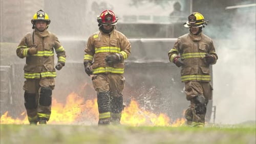 A group of firefighters in full protective gear walks through ground flames during a live fire