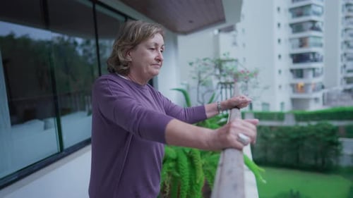 One contemplative senior woman standing at apartment balcony overlooking condominium. A pensive