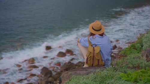 Girl Traveler Sitting on a Rock in a Hat and with a Backpack Enjoying the View of the Ocean