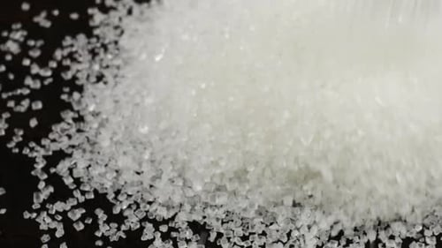 White sugar crystals are sprinkled onto a dark brown wooden surface. A big pile of sugar