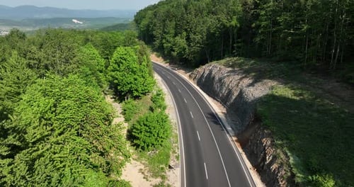 Aerial view of a two-lane road curving through a dense forest. Lush green trees line the road, creat