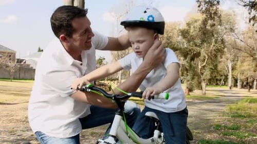 Steadicam Shot Of Father Teaching Son How To Use His His Safety Helmet On A Park Pathway.