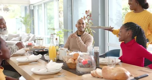 Black woman hearing applause and serving dishes at table while Diverse family welcoming bearded man