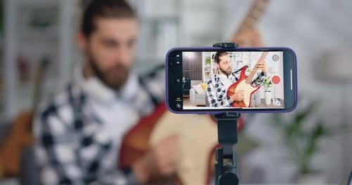 Smartphone con pantalla de cerca. Joven bloguero masculino usa auriculares para tocar la guitarra eléctrica