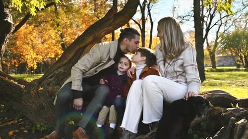 Happy family in an autumn park. Mother, father, son and daughter sitting on a tree trunk, kissing ea