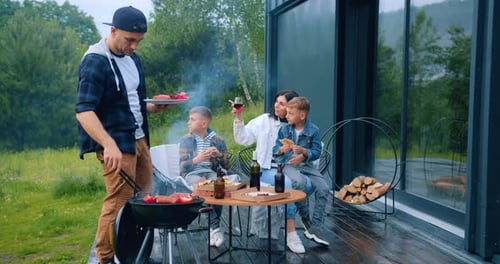 Man cooking vegetables and sausages on grill when children talking with mother around the table.