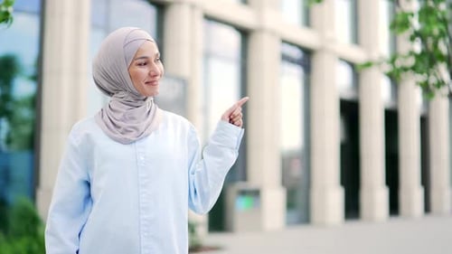 Happy young muslim female employee in hijab pointing fingers at empty space for writing text stand