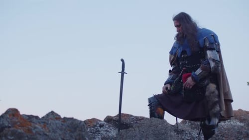 A Man in a Medieval Knight Costume Pulls Out a Sword From a Stone with Both Hands Against the