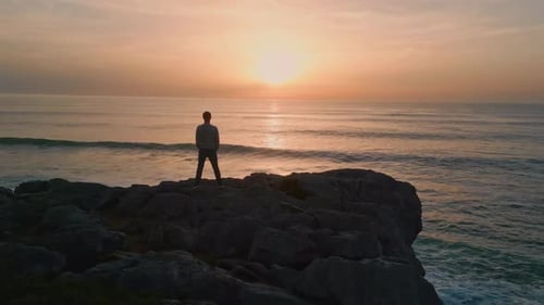 Man Standing on Cliff Watching Golden Sunset Over Rough Ocean Waves